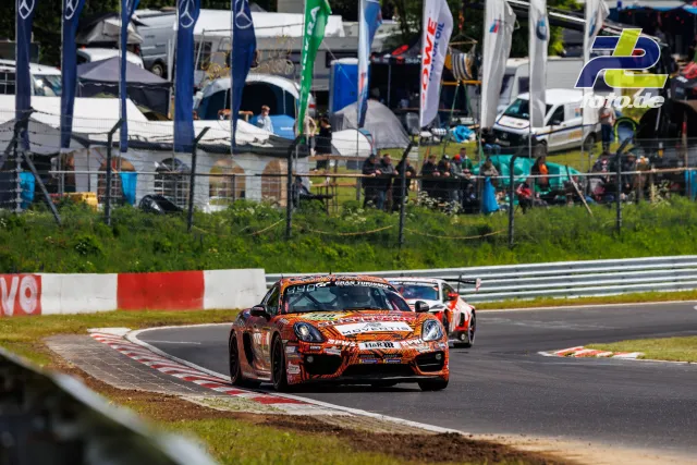 Porsche Cayman GT4 in orangener Lackierung mit Sponsoren auf der Nordschleife, gefolgt von weiteren Fahrzeugen, Fahnenmeer im Fahrerlager.