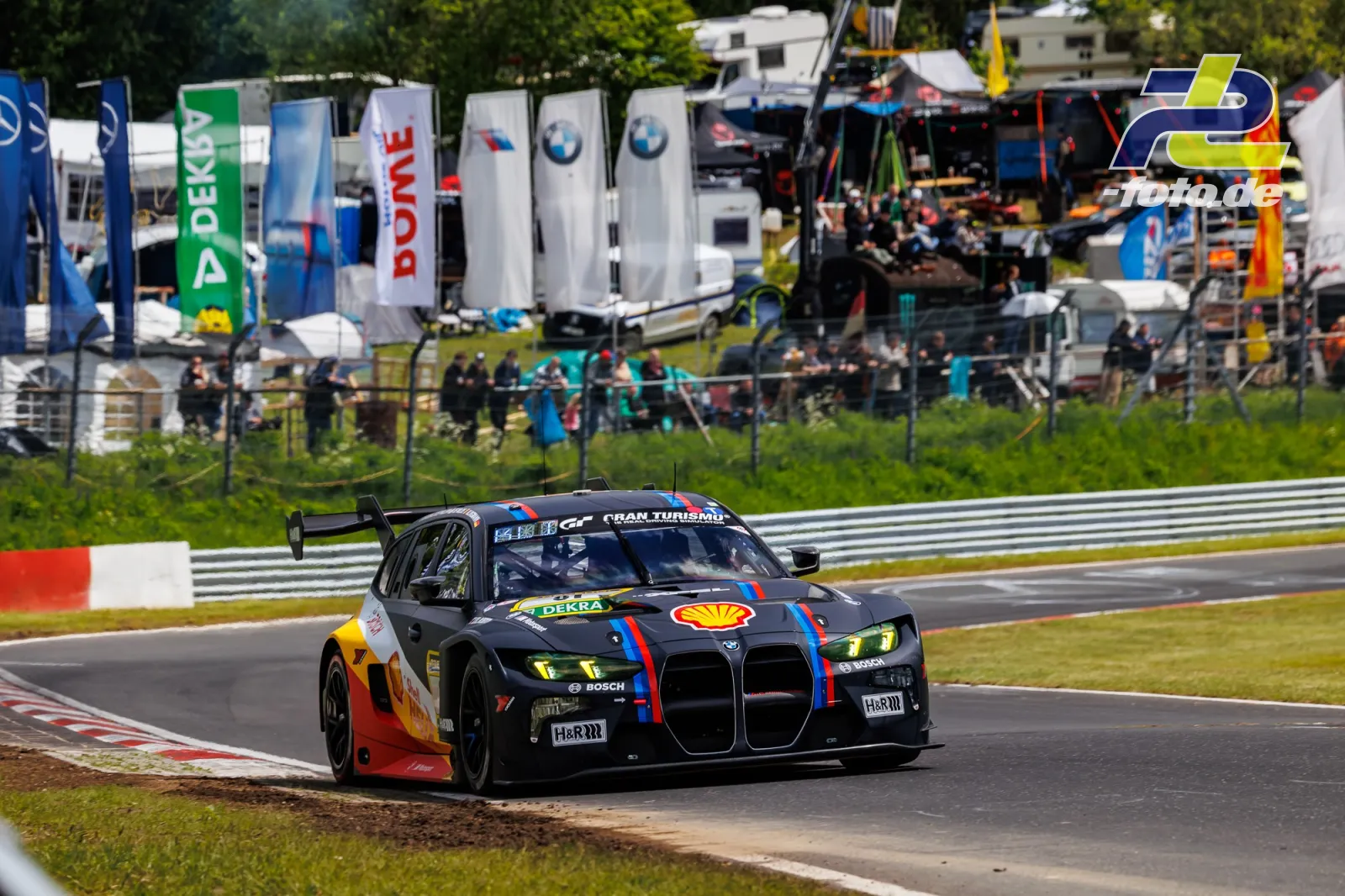 BMW M4 GT3 in Shell-Lackierung auf der Nürburgring-Nordschleife während des Qualifyings zum 24-Stunden-Rennen, im Hintergrund Paddock und Flaggen.