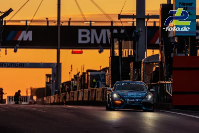 Rennwagen verlässt bei Sonnenuntergang die Boxengasse am Nürburgring, Silhouetten von Boxenteam und BMW-Branding im Hintergrund.