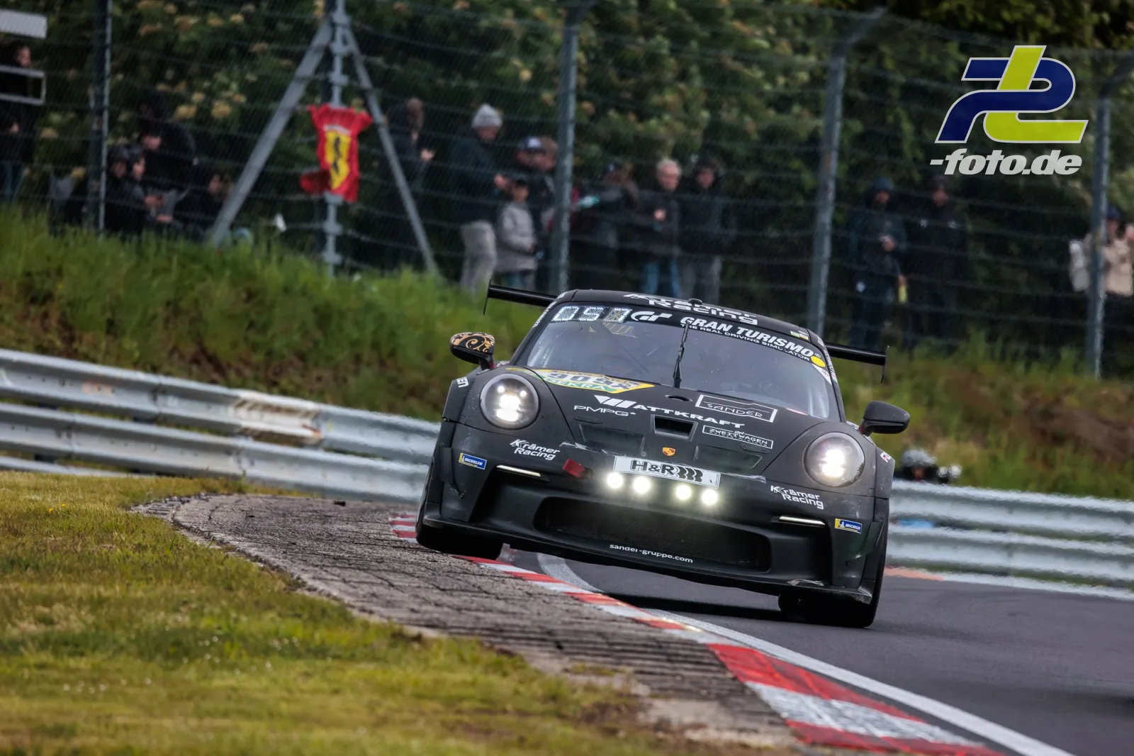 Schwarzer Porsche 911 GT3 R #909 fährt über Randsteine auf der Nürburgring-Nordschleife, Zuschauer hinter Zaun mit Ferrari-Flagge.