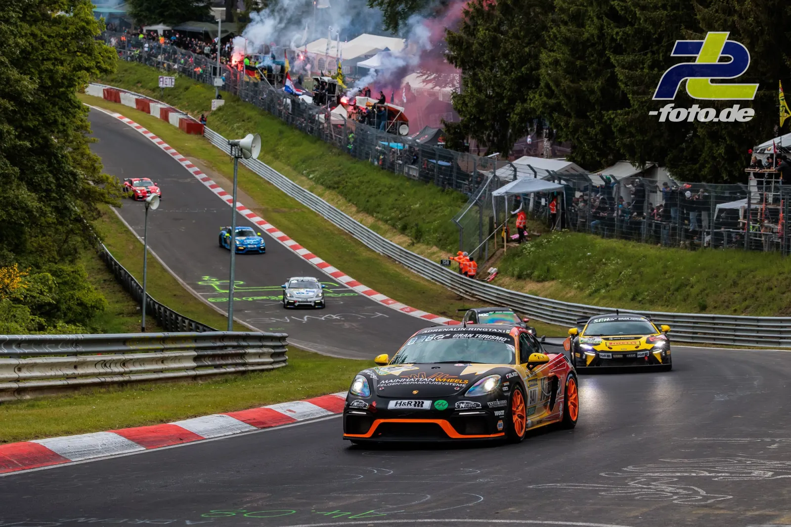Porsche 718 Cayman GT4 Clubsport während der Formationsrunde auf der Nordschleife, gefolgt von mehreren Fahrzeugen. Zuschauer mit Bengalos im Hintergrund.