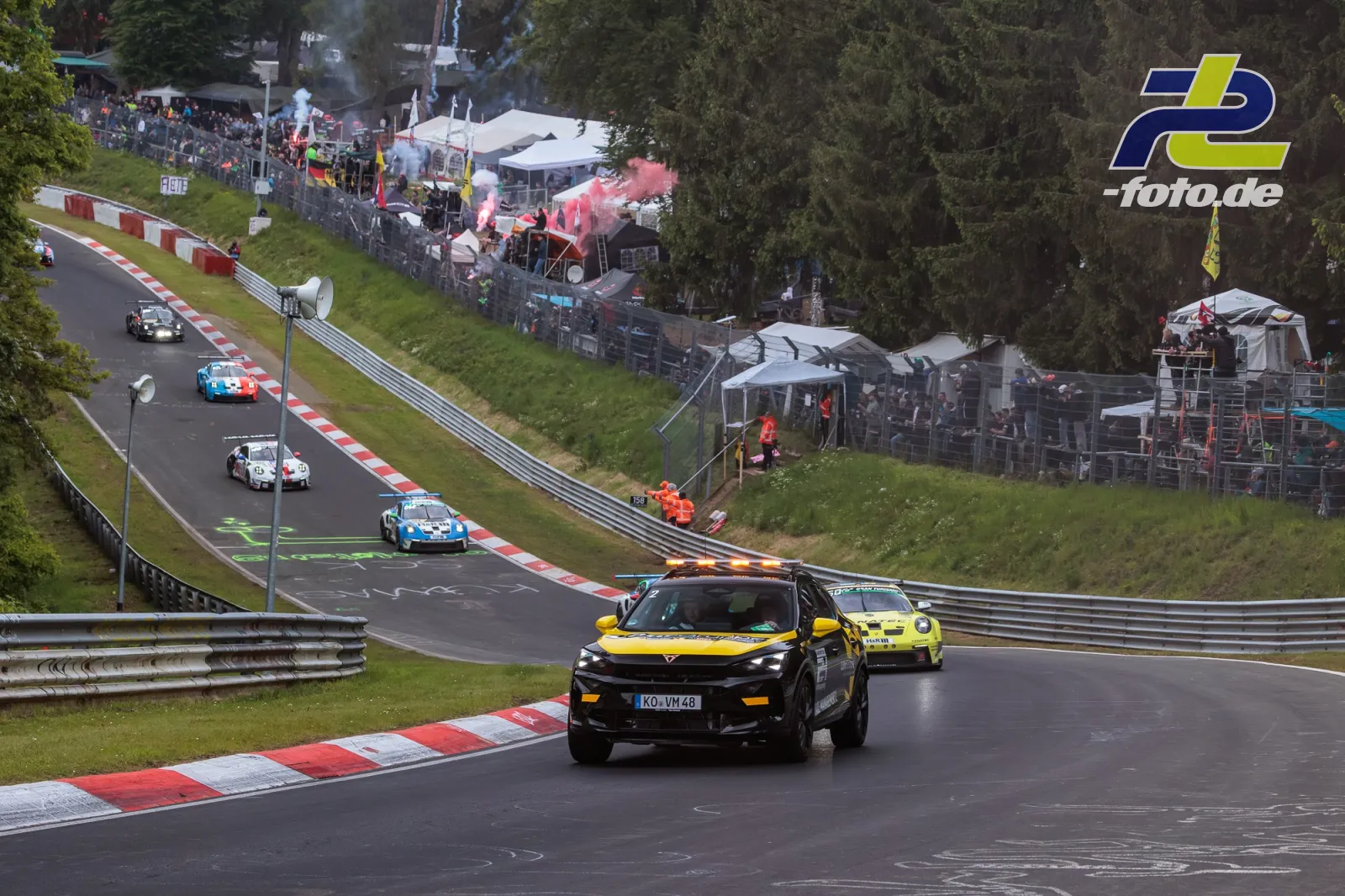 Gelb-schwarzes Fahrzeug führt die Formationsrunde vor Rennwagen auf der Nürburgring-Nordschleife an, Zuschauer und Zelte im Hintergrund.