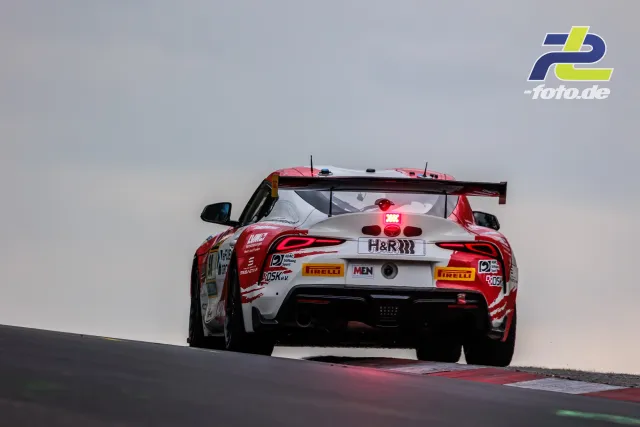 BMW M4 GT3 mit rot-weißer Lackierung fährt über die Kuppe auf dem Nürburgring, Heckansicht mit leuchtenden Bremslichtern.