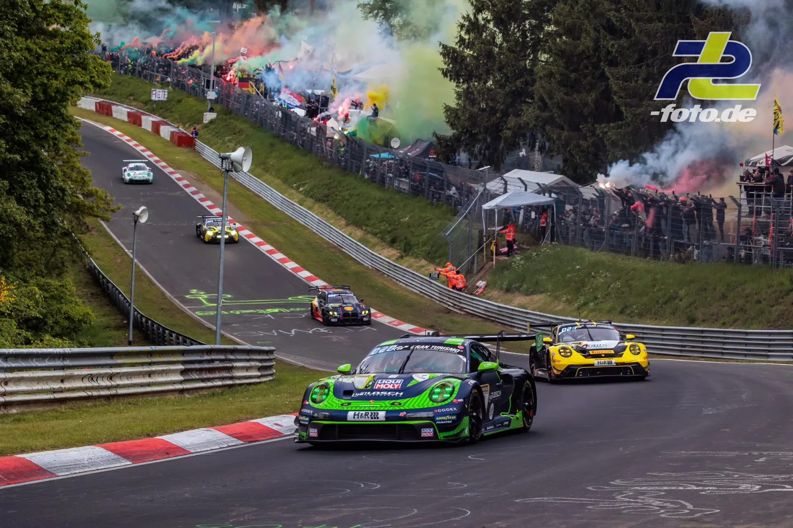 Mehrere Rennwagen in einer Kurve auf der Nordschleife während der Formationsrunde, Zuschauer mit bunten Rauchfackeln am Streckenrand.