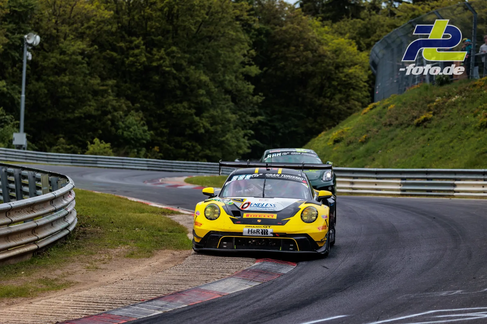 Porsche 911 GT3 R in der Kurve auf der Nürburgring-Nordschleife beim 24h-Rennen, gelbe Lackierung, Zuschauer im Hintergrund.