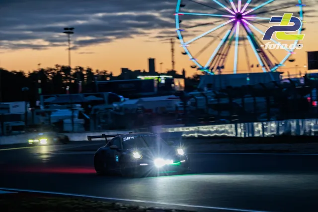 Rennwagen bei Nachtfahrt mit leuchtenden Scheinwerfern vor beleutetem Riesenrad am Nürburgring während des 24h-Rennens.