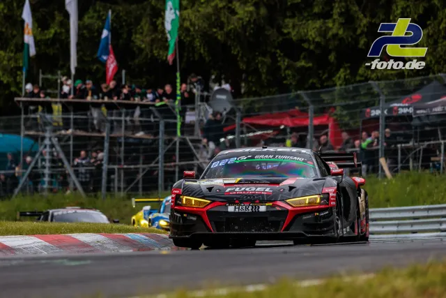 Audi R8 LMS GT3 in schwarz-rot bei der Nordschleife, Zuschauertribünen und Bäume im Hintergrund, vor einem gelb-blauen Verfolger.