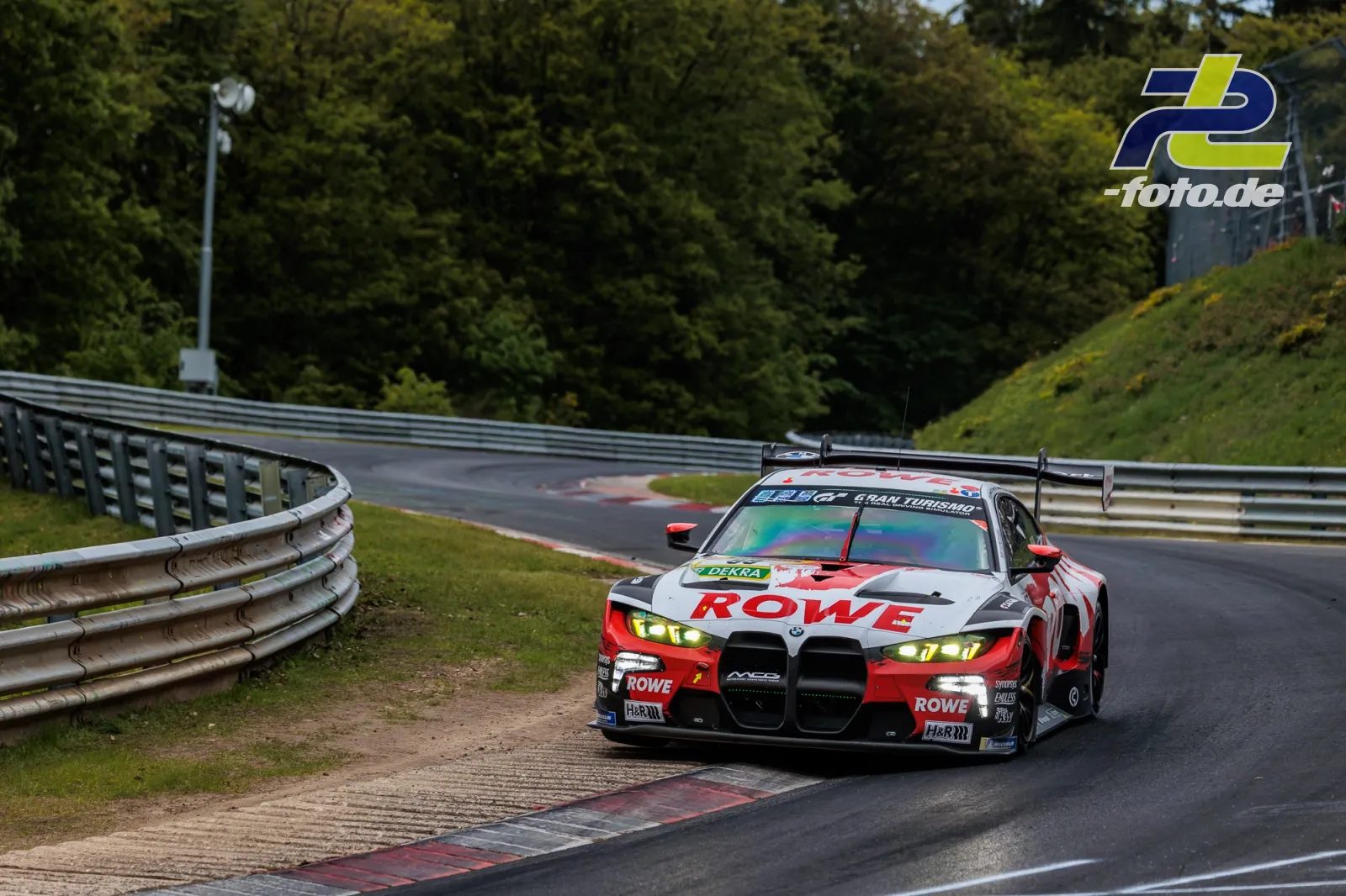 BMW M4 GT3 EVO von ROWE RACING in Kurve auf der Nürburgring-Nordschleife beim 24h-Rennen. Rot-weißes Design mit Heckspoiler.