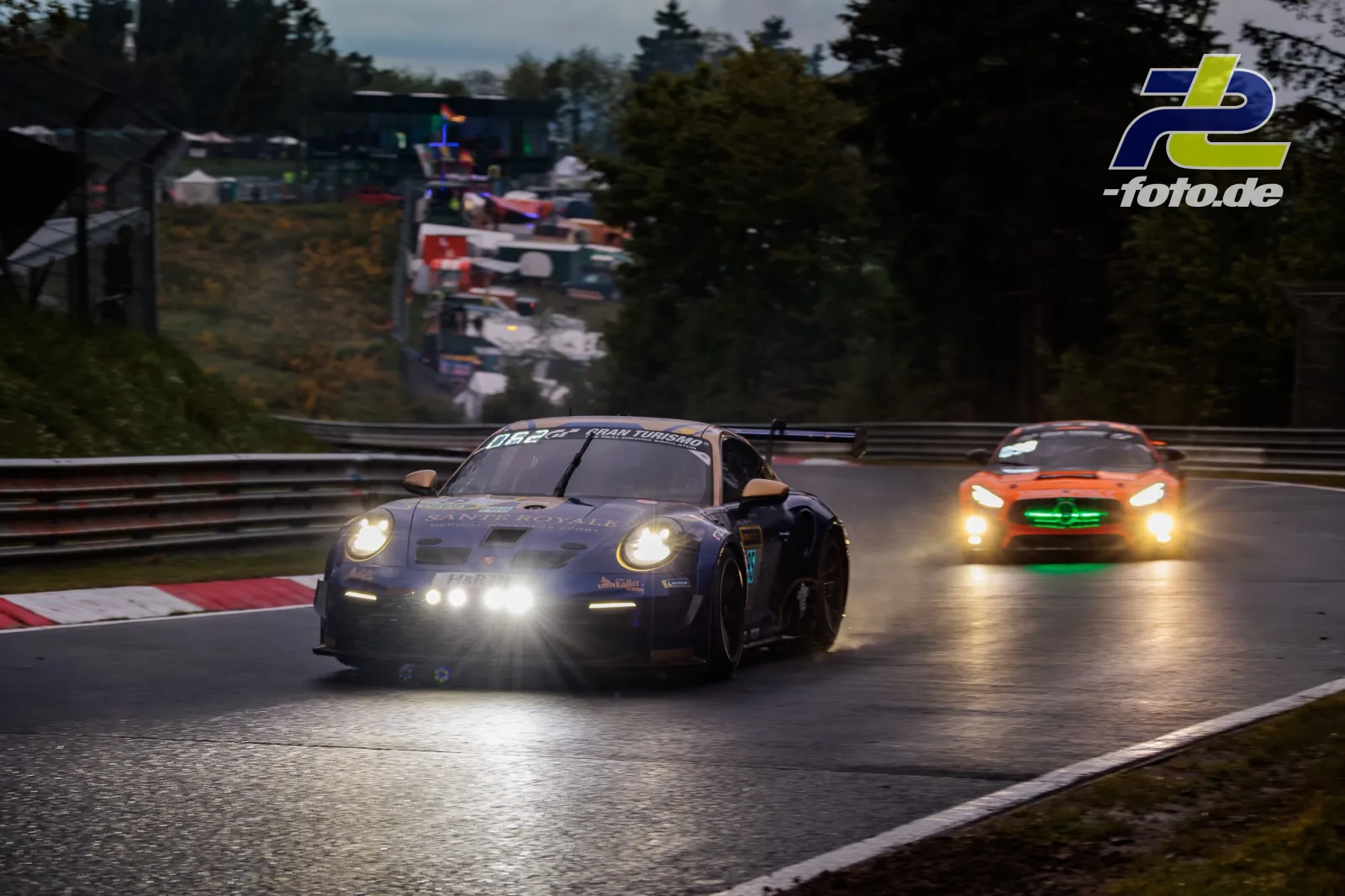 Zwei Rennwagen bei nasser Fahrbahn auf der Nordschleife, das führende Fahrzeug ist ein dunkler Porsche, gefolgt von einem orangefarbenen GT-Sportwagen.