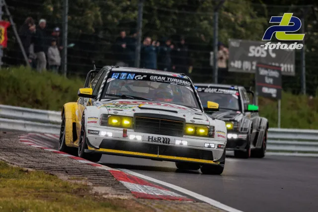 Mercedes-Benz 190 E in weiß-gelber Lackierung auf der Nordschleife beim 24h-Rennen, gefolgt von einem grauen Fahrzeug, Zuschauer im Hintergrund.
