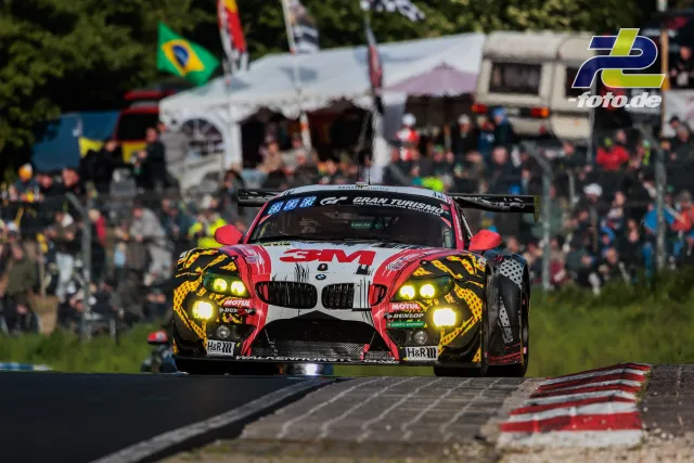 BMW mit rot-gelb-schwarzer Lackierung auf der Nordschleife beim 24h-Rennen, Zuschauer und Flaggen im Hintergrund