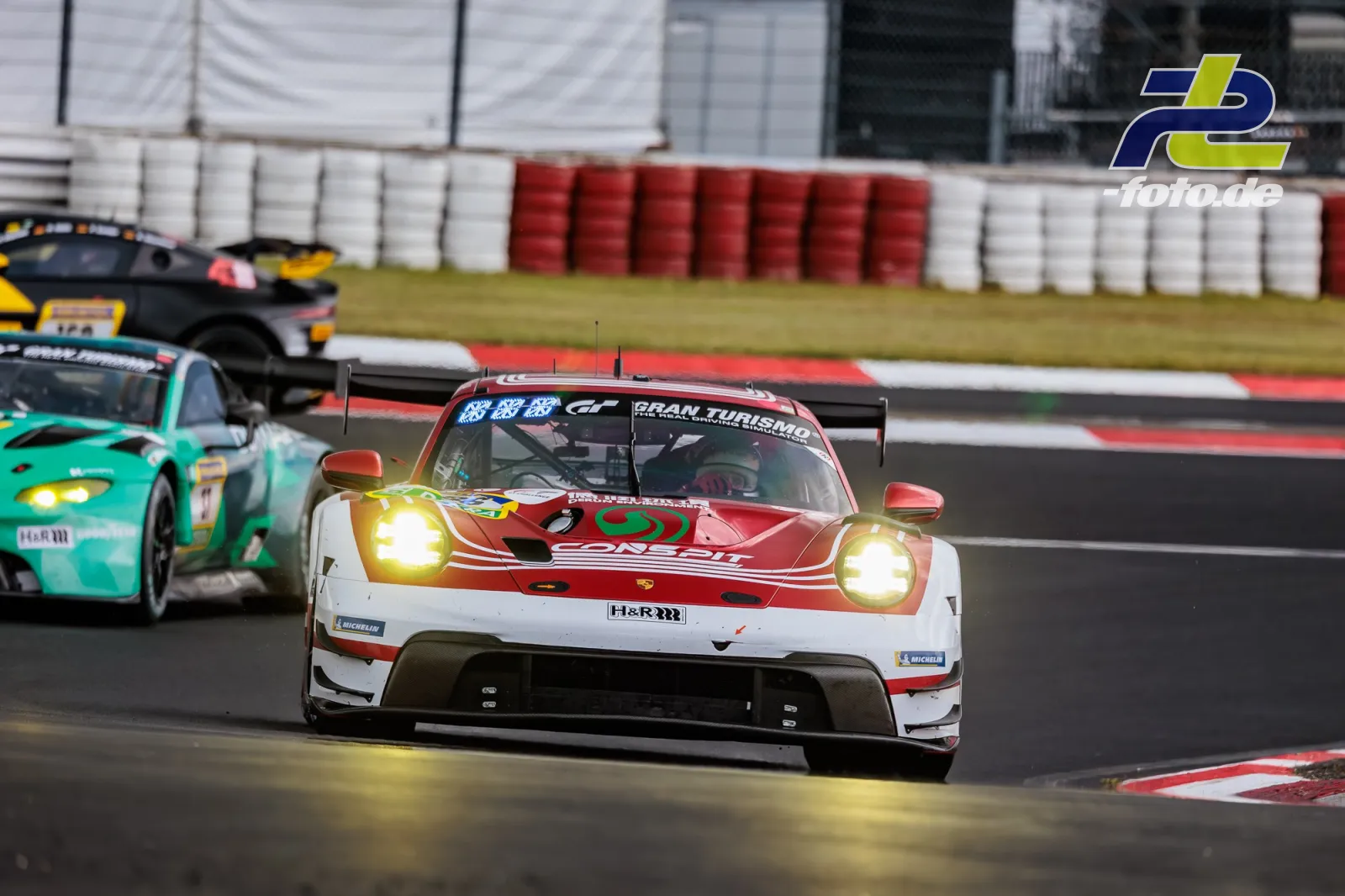 Porsche 911 GT3 R in Rot-Weiß während Qualifying 1 auf dem Nürburgring, verfolgt von einem türkisfarbenen Fahrzeug. Scheinwerfer eingeschaltet.