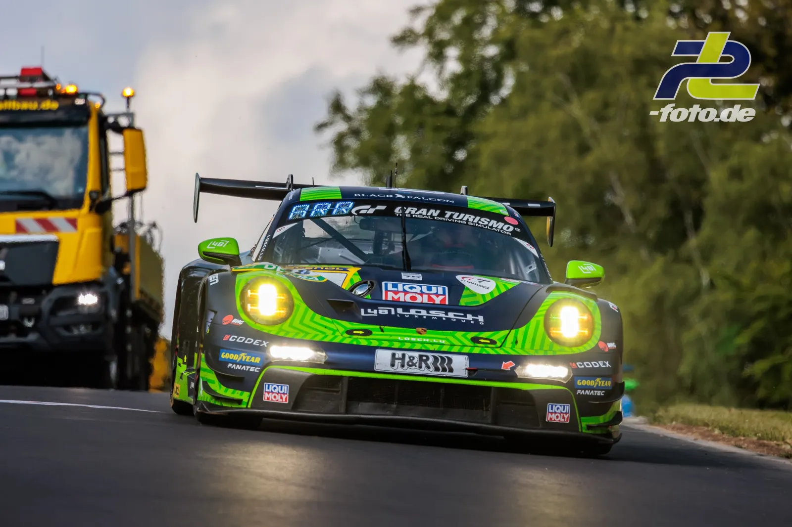 Porsche 911 GT3 R in neongrüner Lackierung mit LIQUI MOLY Sponsoring passiert Bergungsfahrzeug auf der Nordschleife bei Tageslicht.