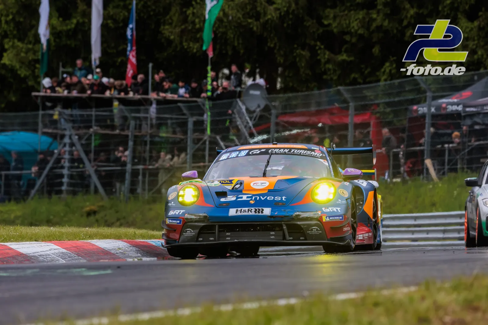 Porsche 911 GT3 R in blau-orange Lackierung mit gelben Scheinwerfern auf dem Nürburgring, Zuschauertribüne im Hintergrund.