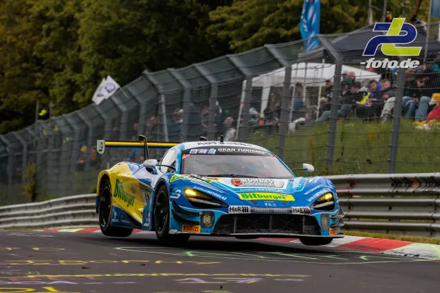 McLaren 720S GT3 mit blau-gelber Bitburger-Lackierung während des 24h-Rennens auf der Nürburgring-Nordschleife vor Zuschauertribüne.