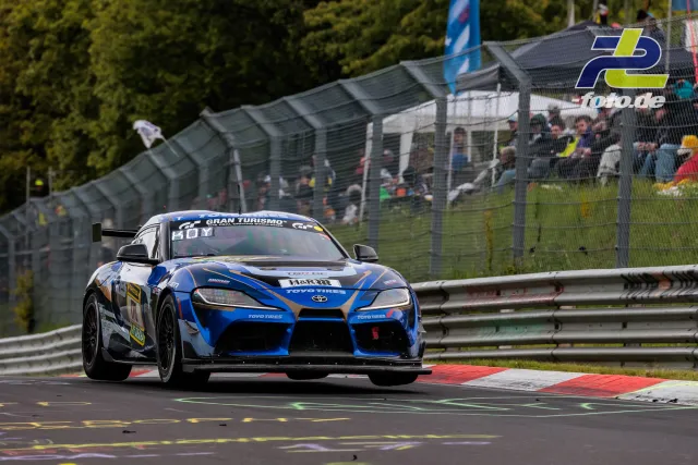 Toyota GR Supra #170 in blau-schwarz auf der Nordschleife beim 24h-Rennen, Zuschauer und Tribüne im Hintergrund.