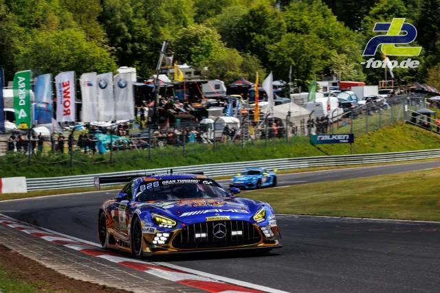Mercedes-AMG GT3 in blau-orange auf der Nürburgring-Nordschleife, im Hintergrund Boxenbereich mit Zuschauern.