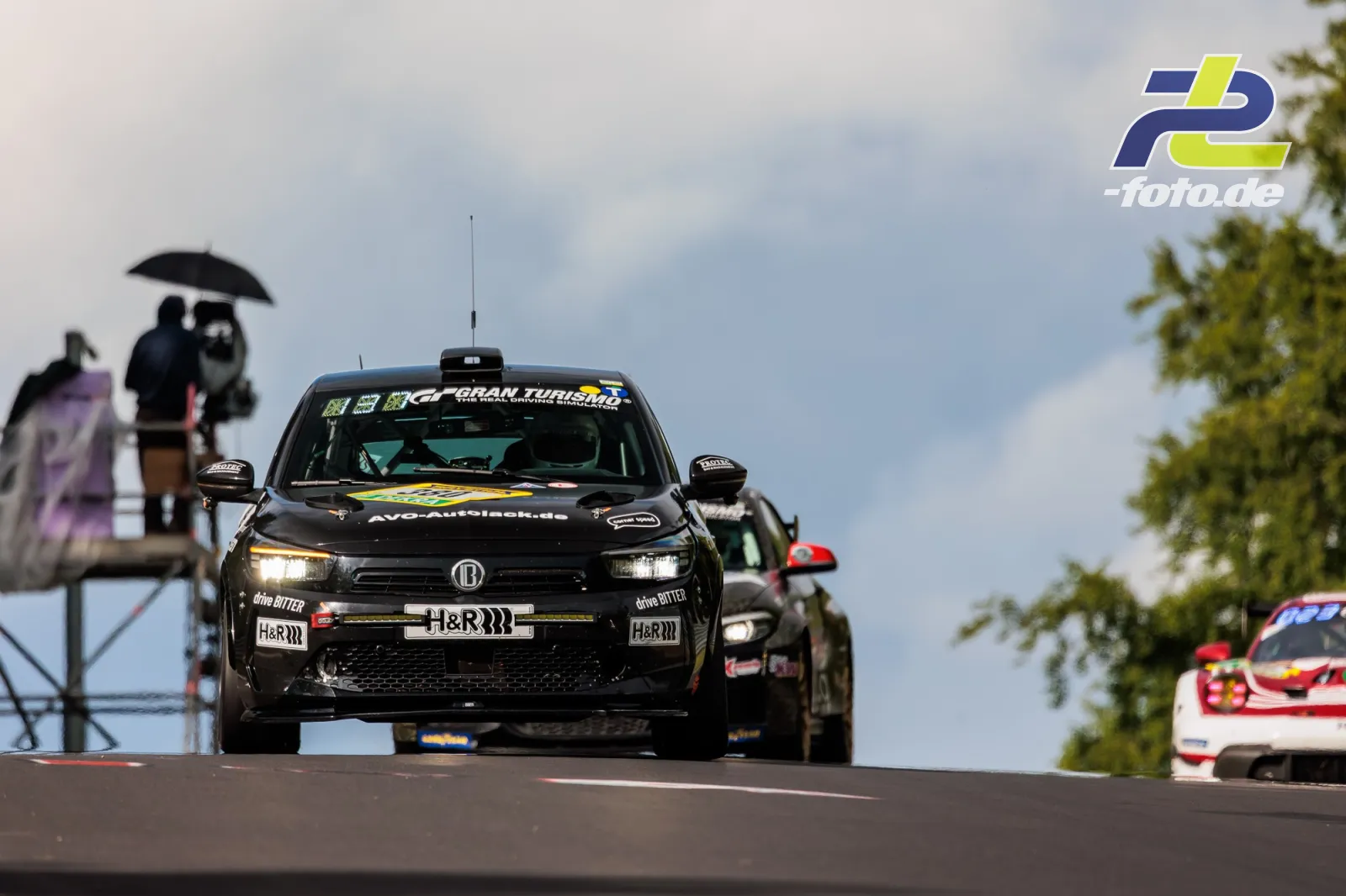 Schwarzer Opel Corsa GS Line 130* bei der Fahrt über eine Kuppe beim 24h-Rennen am Nürburgring, im Hintergrund Streckenposten mit Schirm.