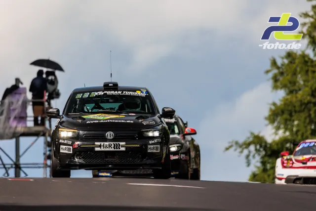 Schwarzer Opel Astra fährt über die Kuppe auf der Nordschleife beim 24h-Rennen, Streckenposten und weitere Fahrzeuge im Hintergrund.