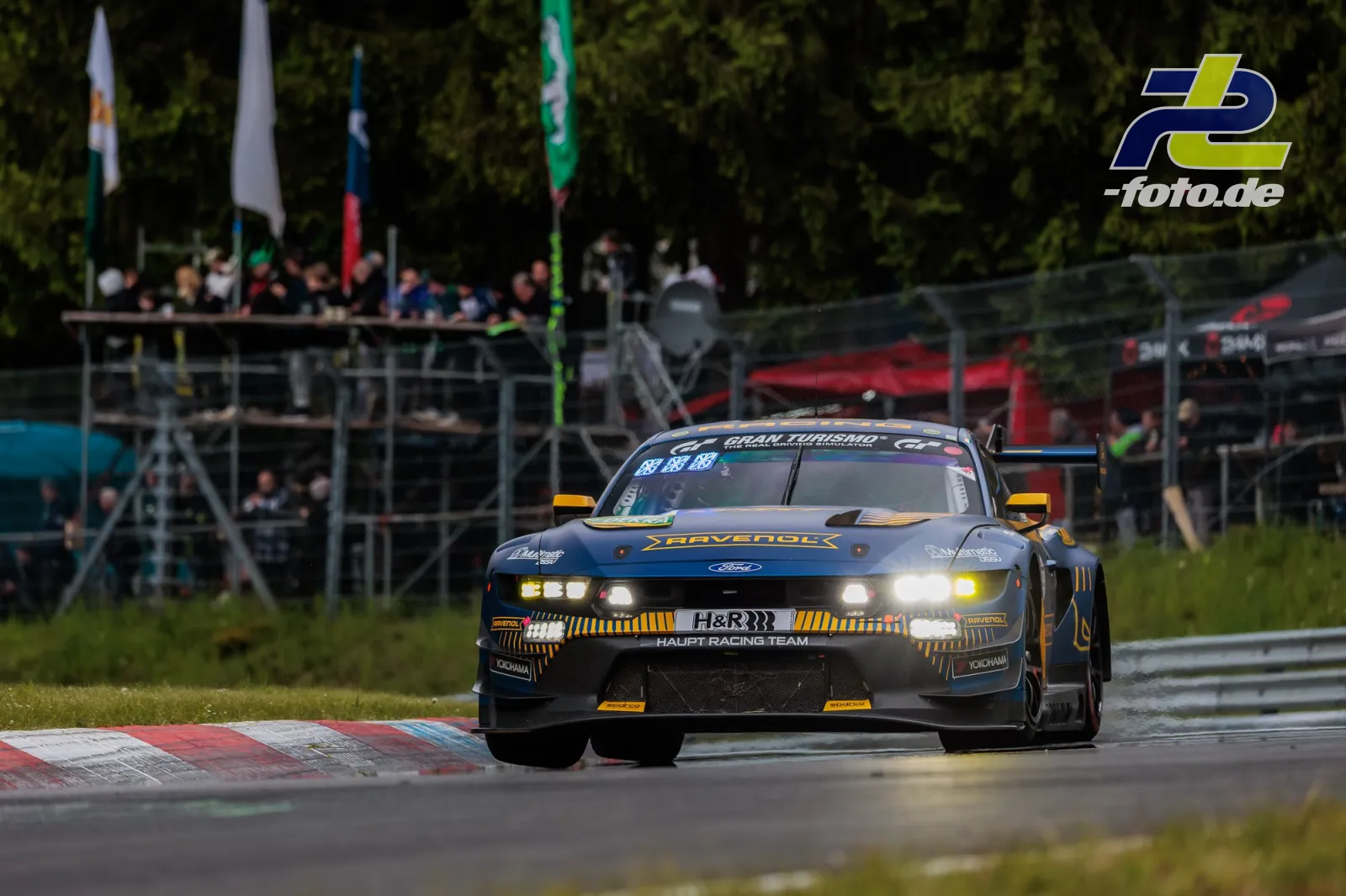 Ford Mustang GT mit Ravenol-Lackierung auf der Nordschleife, Zuschauer auf Tribüne im Hintergrund, Randsteinberührung.