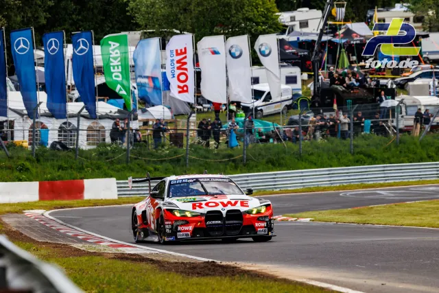 BMW M4 GT3 von ROWE Racing im Qualifying auf der Nürburgring-Nordschleife, Zuschauerbereich mit Sponsorenfahnen im Hintergrund