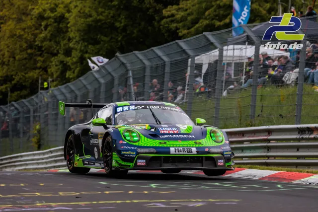 Porsche 911 GT3 R #48 in grün-blauer Lackierung fährt durch eine Kurve auf dem Nürburgring, Zuschauertribüne im Hintergrund.