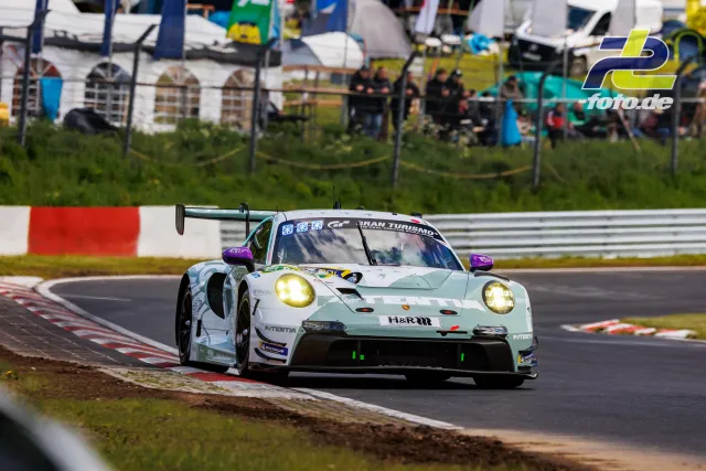 Porsche 911 GT3 R in mintgrün mit violetten Spiegelkappen auf der Nordschleife, Gelbe Scheinwerfer eingeschaltet, Zuschauer im Hintergrund.