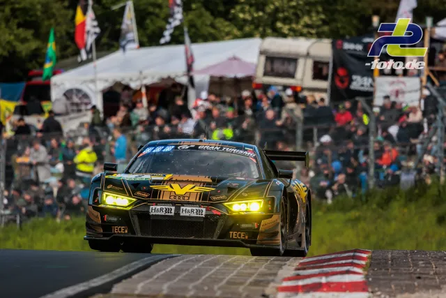 Audi R8 LMS GT3 mit leuchtenden Scheinwerfern auf der Nordschleife des Nürburgrings vor Zuschauertribünen.