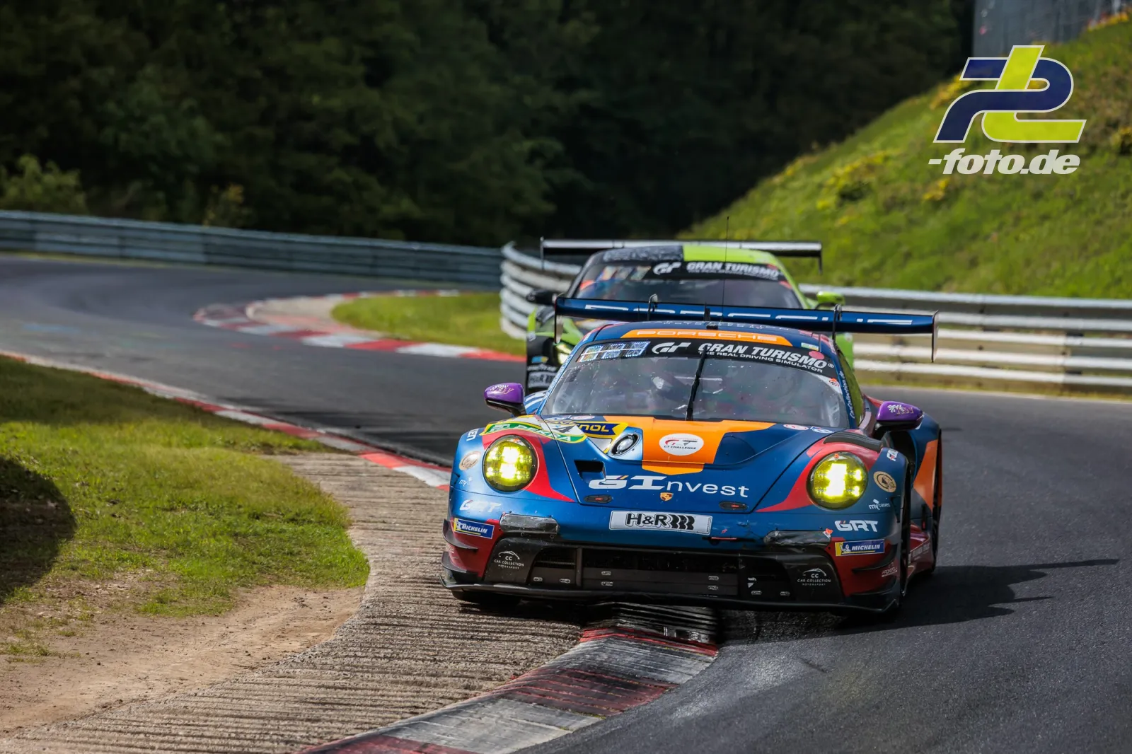 Porsche 911 GT3 R in blau-orange mit GT Invest Lackierung überfährt Randstein auf der Nürburgring-Nordschleife, gefolgt von einem weiteren GT3-Fahrzeug.
