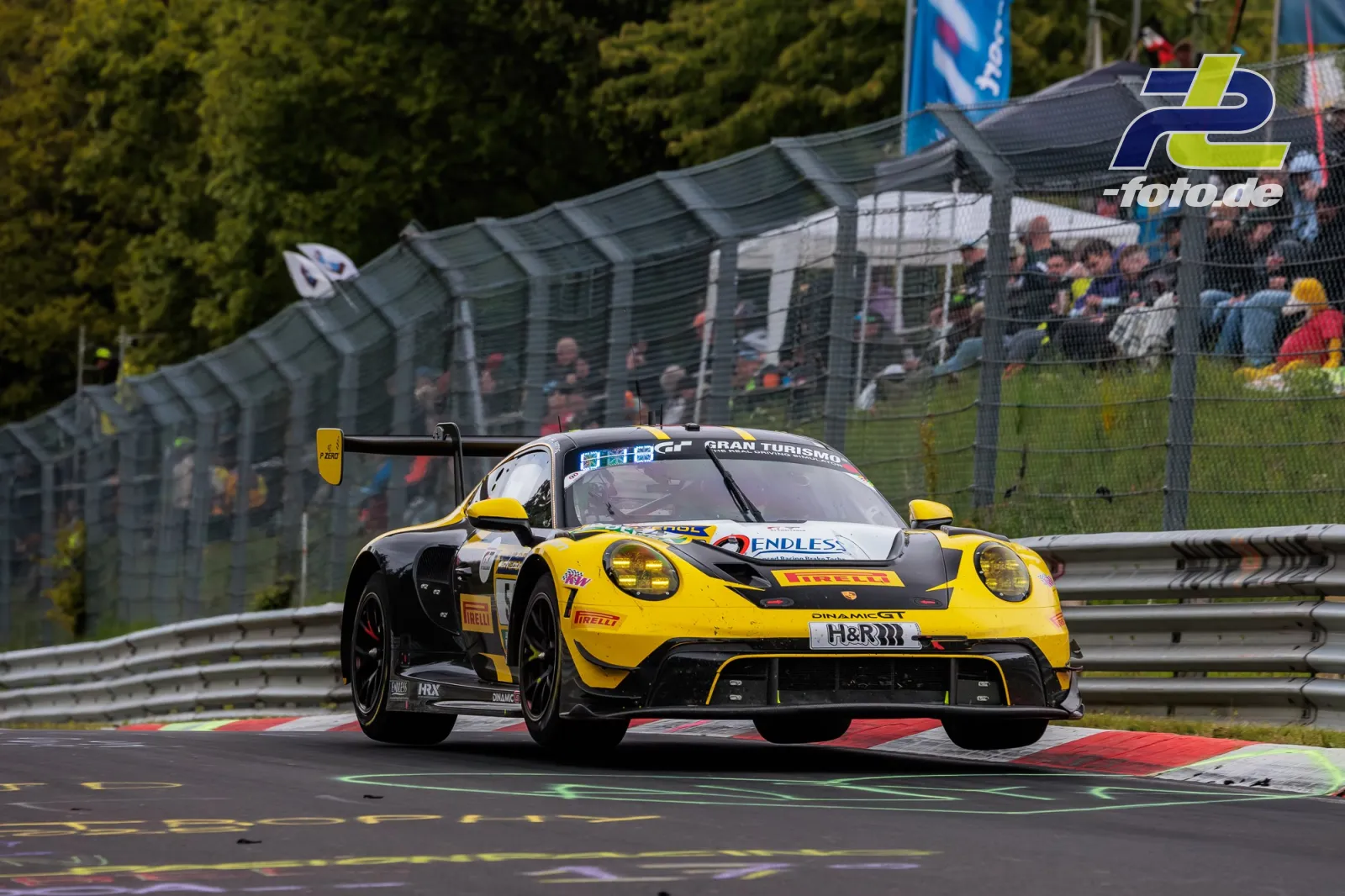 Gelb-schwarzer Porsche 911 GT3 R von Dinamic GT beim 24h-Rennen am Nürburgring mit Zuschauertribüne im Hintergrund.