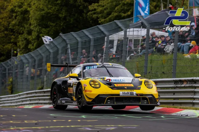 Porsche 911 GT3 R in gelb-schwarz auf der Nordschleife während des 24h-Rennens, Zuschauertribüne im Hintergrund.