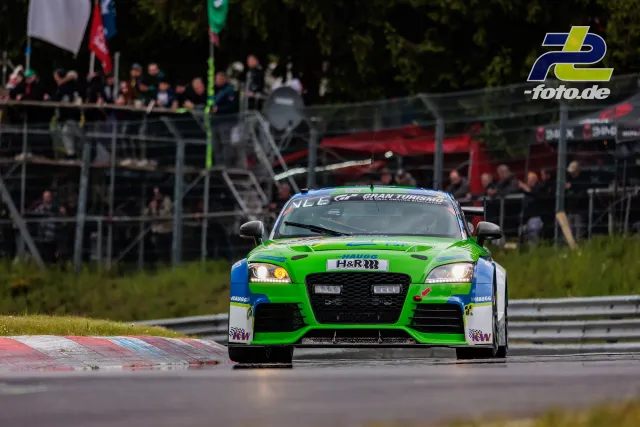 Audi TT RS in giftgrün-blauer Lackierung frontal auf der Nordschleife, Zuschauertribüne im Hintergrund.