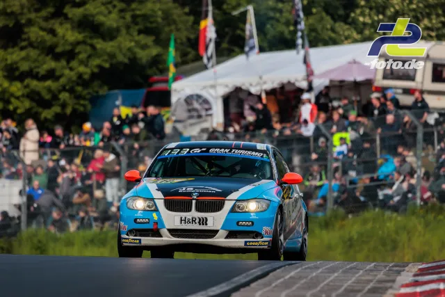 BMW mit blau-weißer Lackierung in Kurve am Nürburgring, Zuschauer und Zelte im Hintergrund während 24h-Rennen.