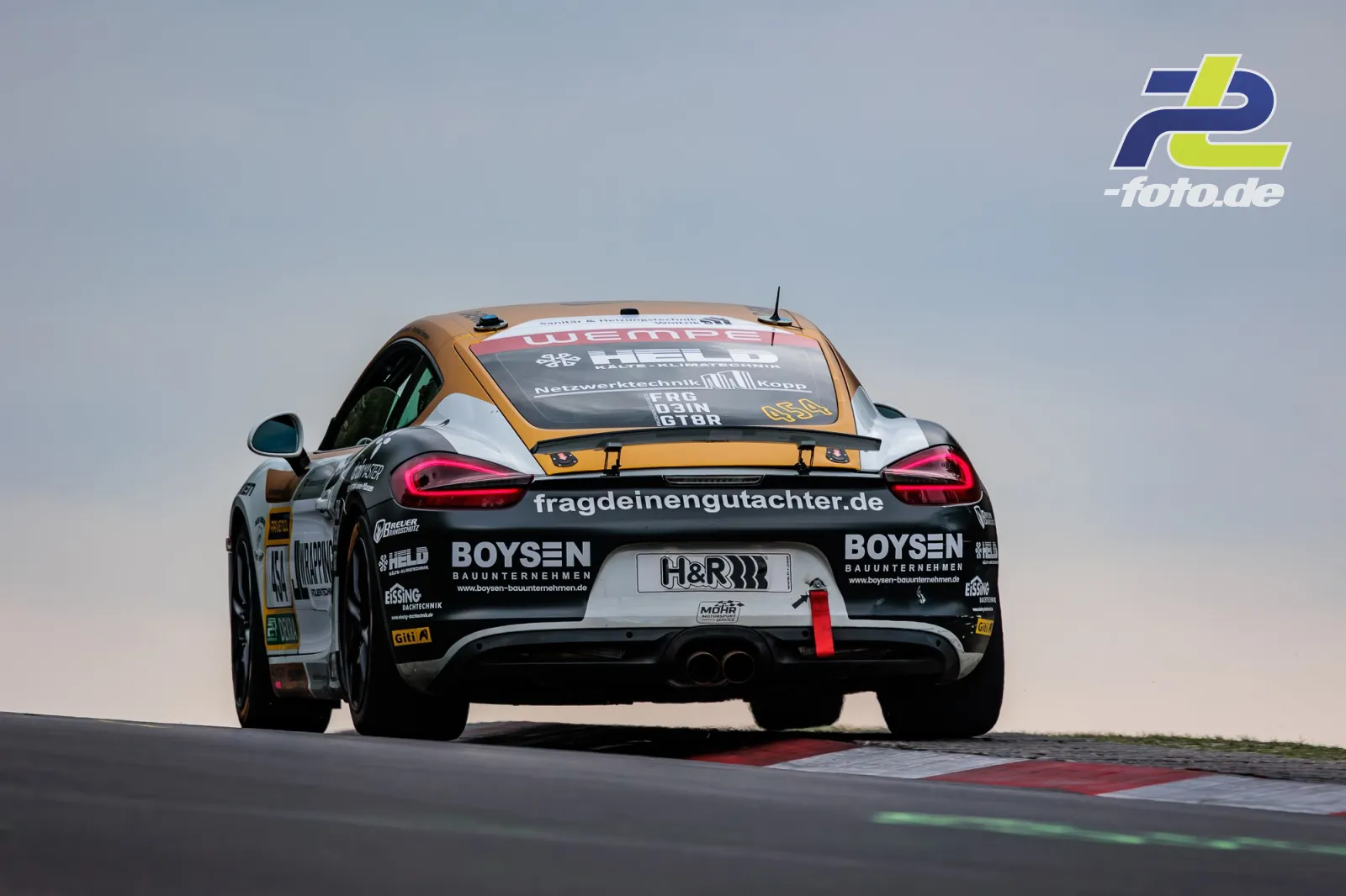 Porsche Cayman CM12 Startnummer 454 von hinten beim 24h-Rennen auf dem Nürburgring, Fahrzeug fährt über eine Kuppe mit Randsteinen.