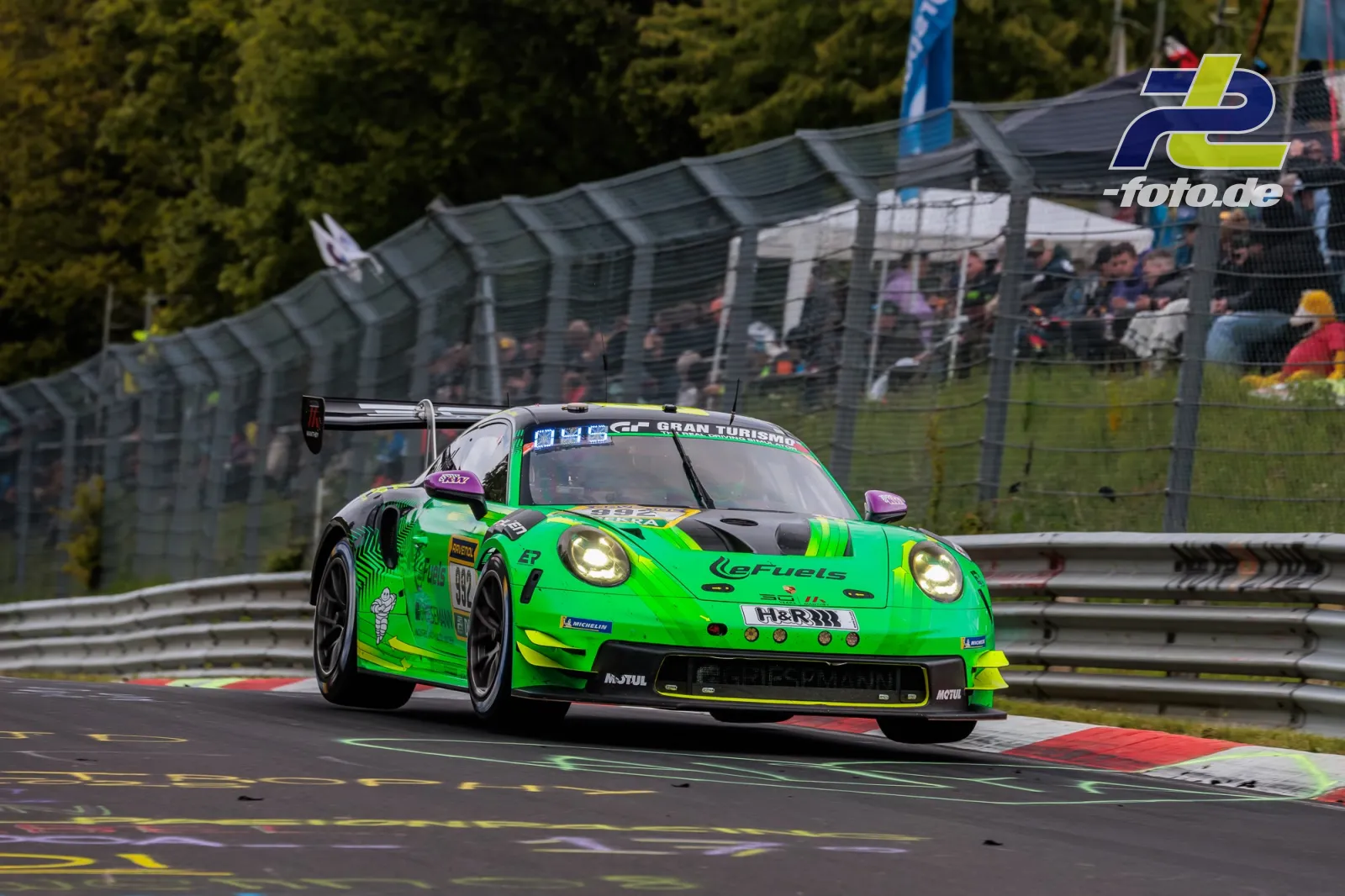 Grüner Porsche 911 GT3 R in einer Kurve auf der Nürburgring-Nordschleife während des 24-Stunden-Rennens, Zuschauertribüne im Hintergrund.