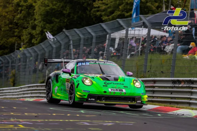Grüner Porsche 911 GT3 R in einer Kurve auf der Nürburgring-Nordschleife während des 24-Stunden-Rennens, Zuschauertribüne im Hintergrund.