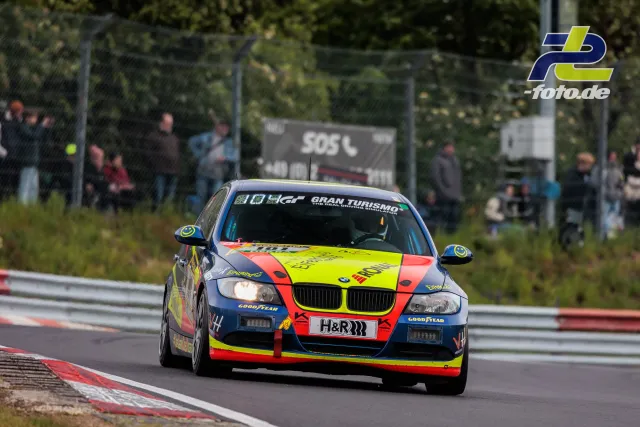 Bunter BMW 3er mit neongelb-rot-blauer Lackierung auf der Nordschleife während des 24h-Rennens, Zuschauer im Hintergrund.