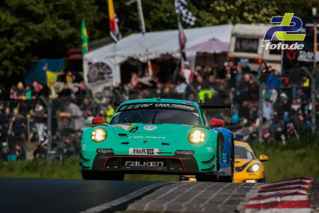Türkiser Porsche 911 GT3 R Nr. 44 mit Falken-Lackierung führt im Rennen vor gelbem Verfolger, Zuschauer und Fahrerlager im Hintergrund.