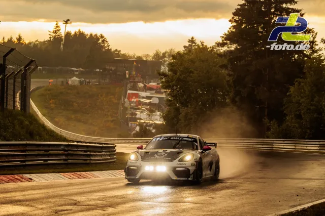 Porsche 718 Cayman GT4 im Gegenlicht auf nasser Nordschleife bei Sonnenuntergang, Gischt sprüht von der Fahrbahn.