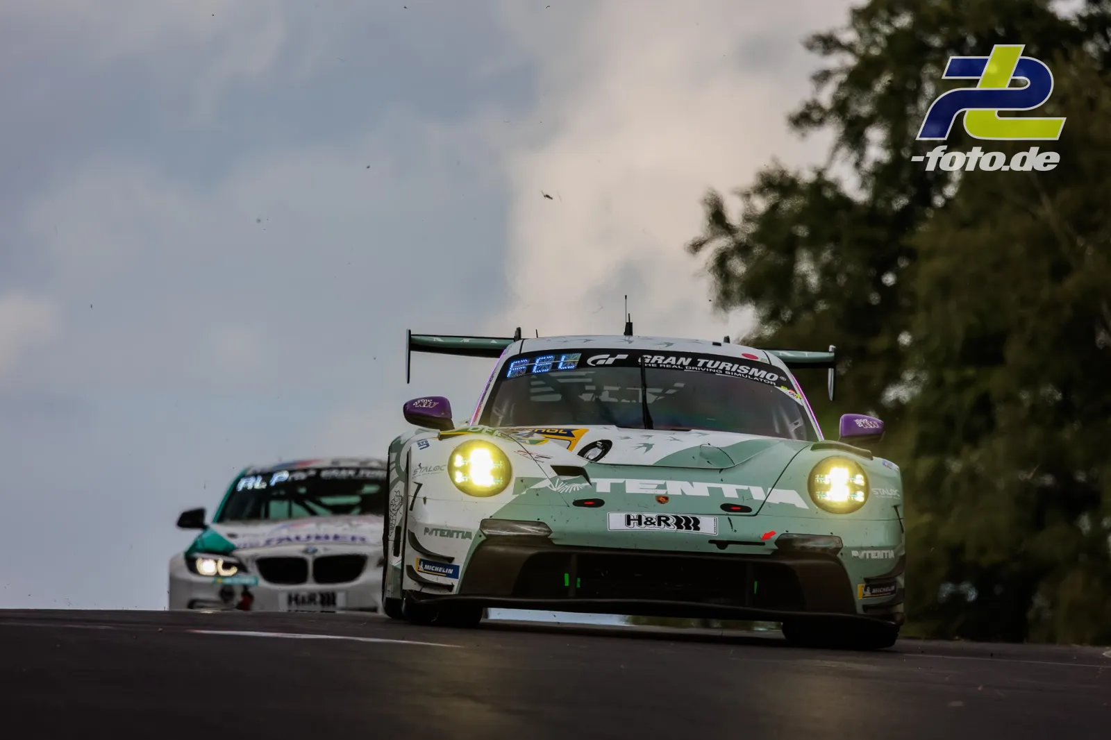 Porsche 911 GT3 R mit mintgrün-weißer Lackierung verfolgt von einem BMW während 24h Nürburgring Rennen bei Dämmerung mit eingeschalteten Scheinwerfern.
