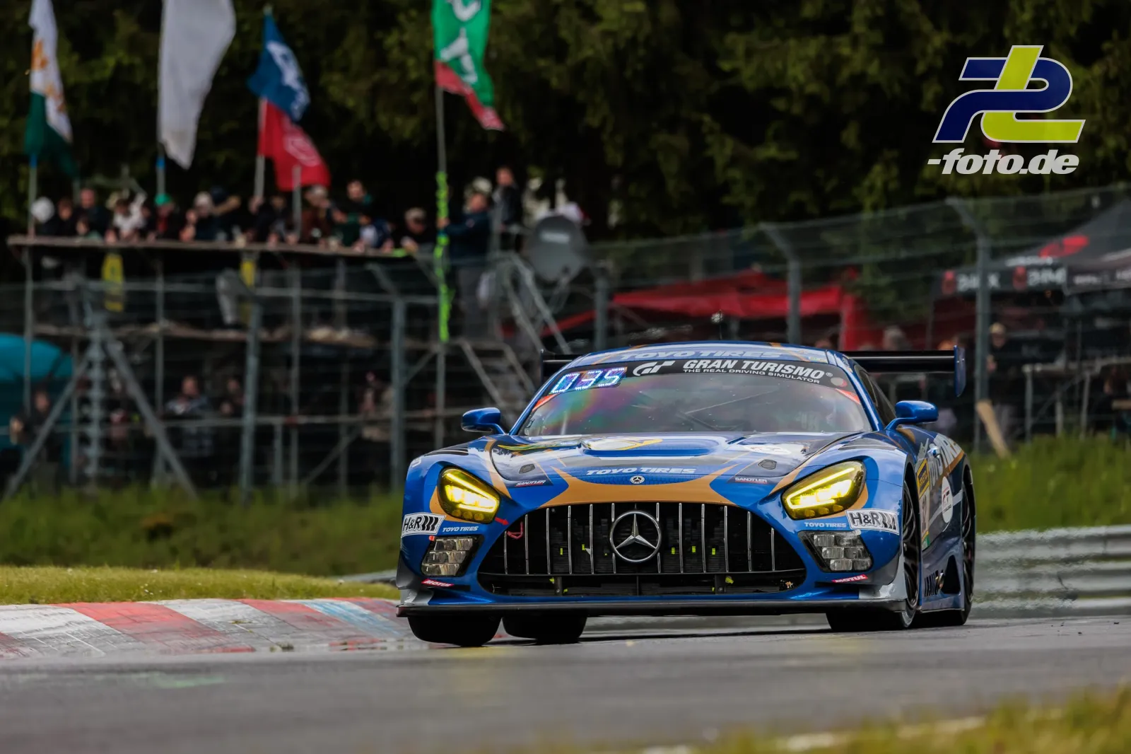 Mercedes-AMG GT3 in blau-oranger Lackierung während des 24h-Rennens auf der Nürburgring-Nordschleife vor Tribüne.
