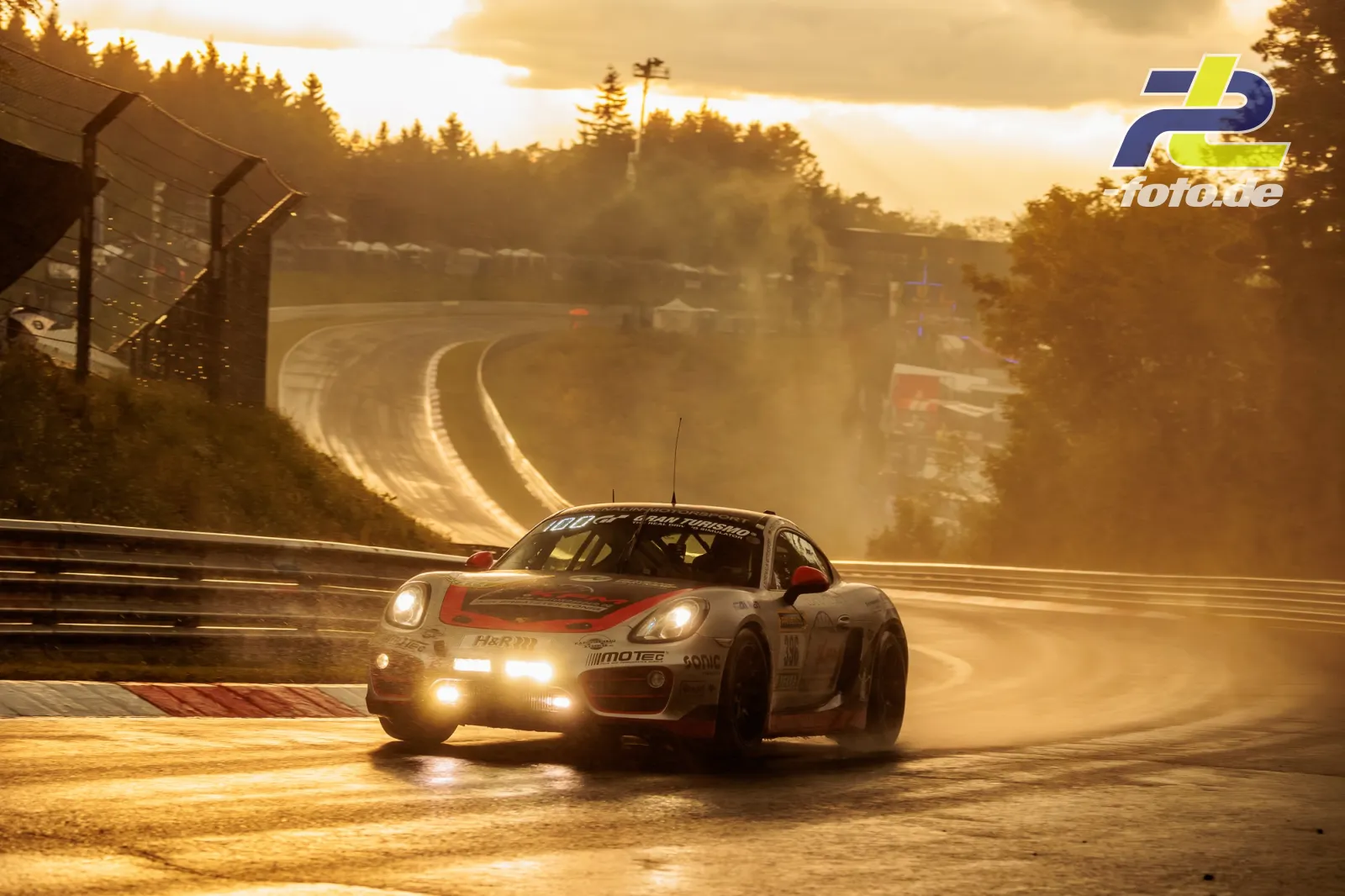 Porsche Cayman S bei Gegenlicht im Qualifying zum 24h-Rennen Nürburgring. Das Fahrzeug fährt durch eine Kurve bei nasser Strecke.