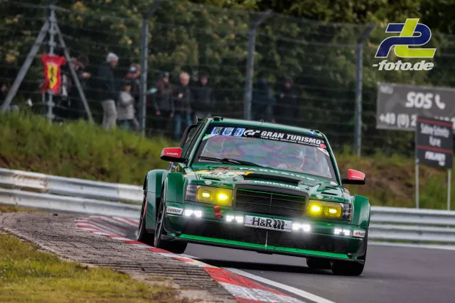 Grüner Mercedes-Benz 190E mit gelben Scheinwerfern auf der Nordschleife beim 24h-Rennen, Zuschauer im Hintergrund.