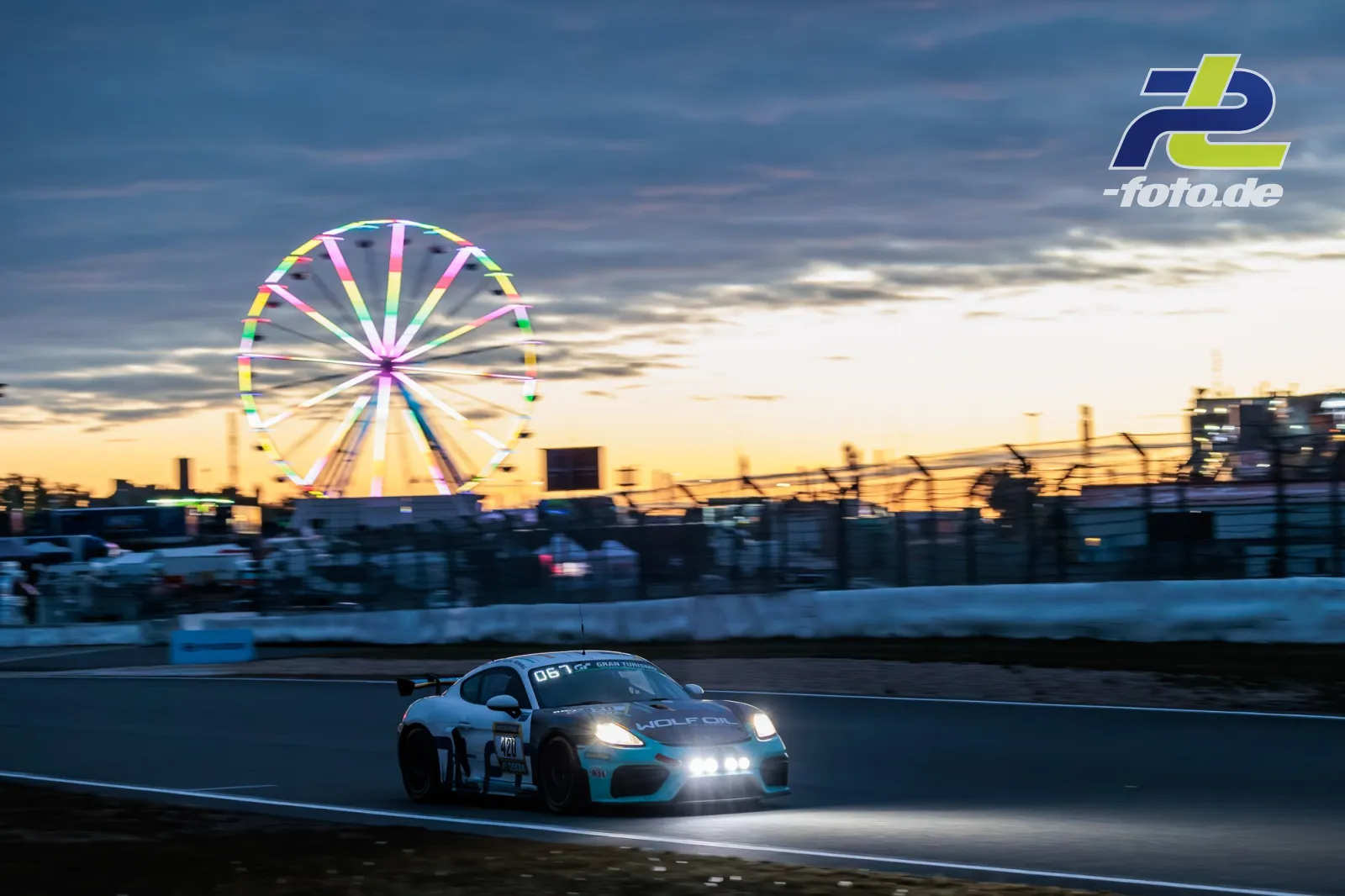 Porsche 718 Cayman GT4 mit Startnummer 420 bei Dämmerung auf dem Nürburgring, Riesenrad im Hintergrund während des 24h-Rennens.