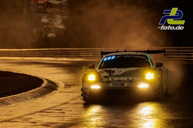 Porsche 911 GT3 R mit leuchtenden Scheinwerfern fährt bei Nässe durch eine Kurve am Nürburgring während des Qualifyings zur Nacht.