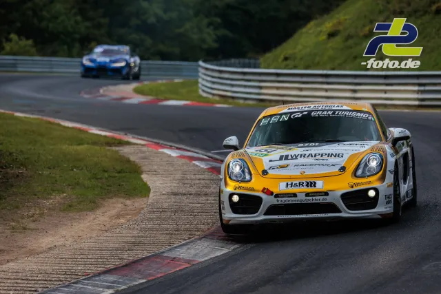 Porsche Cayman in orange-weißer Lackierung in Kurve auf der Nordschleife, BMW im Hintergrund, 24h Nürburgring Rennen.