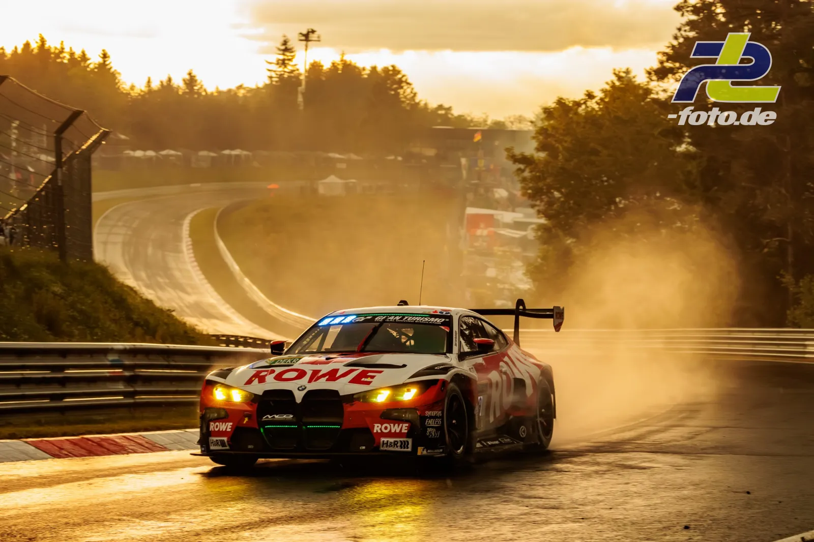 BMW M4 GT3 EVO von ROWE RACING im Gegenlicht auf nasser Strecke beim 24h-Rennen Nürburgring Qualifying, Kurvenausgang mit Wald im Hintergrund.