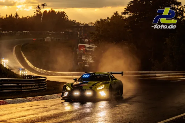 GT-Rennwagen fährt bei Dämmerung und nasser Strecke durch eine Kurve auf dem Nürburgring, eingeschaltete Scheinwerfer, stimmungsvolles Gegenlicht.