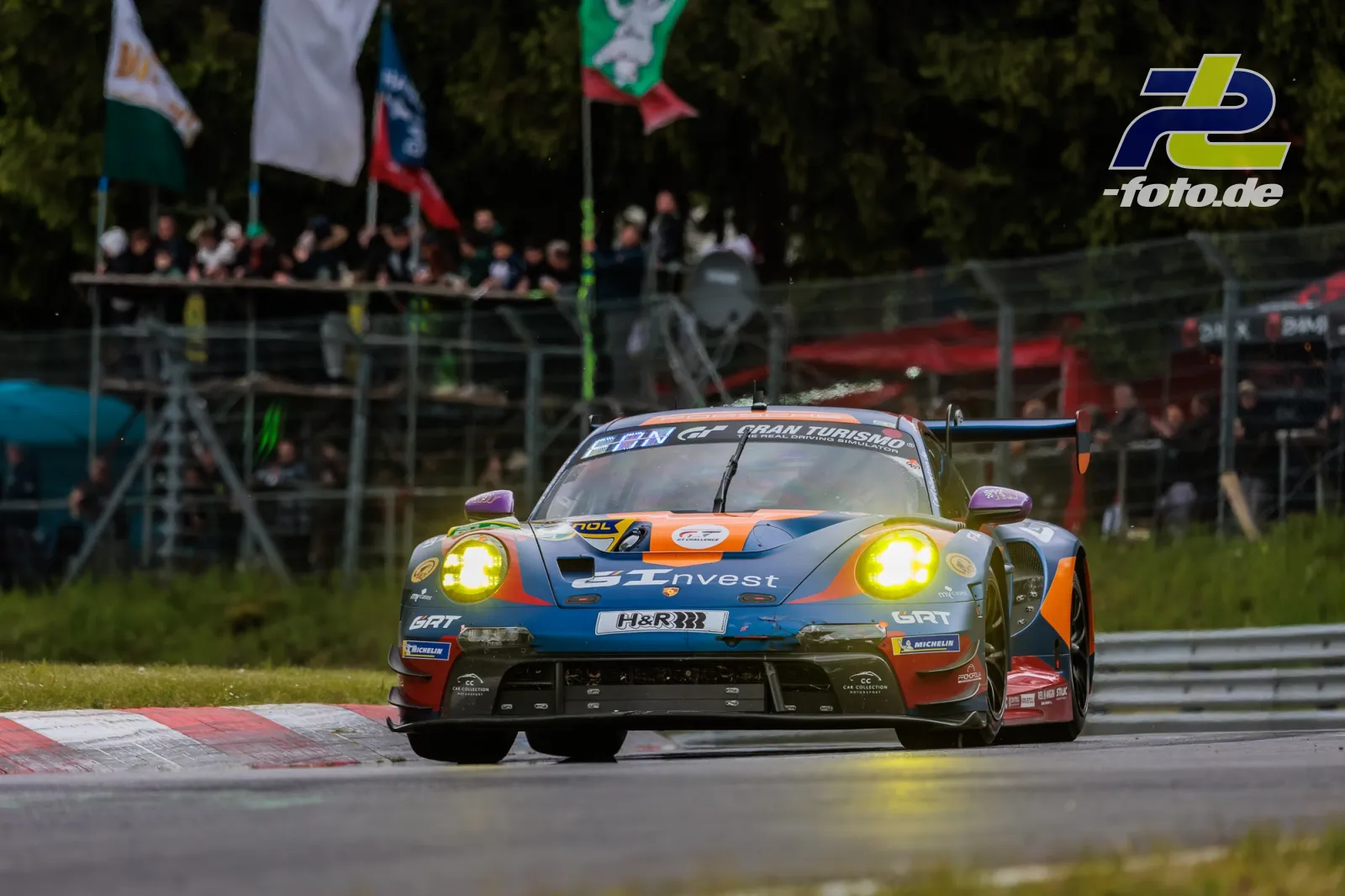 Porsche 911 GT3 R im Gulf-Design auf der Nordschleife beim 24h-Rennen mit eingeschalteten Scheinwerfern vor Zuschauertribüne.