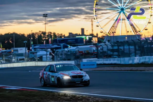 BMW Nummer 108 im Abendhimmel auf dem Nürburgring, im Hintergrund leuchtet ein Riesenrad bei Sonnenuntergang während des 24h-Rennens.